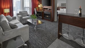 A modern hotel room with a sofa, armchair, coffee table, TV, fireplace, and a dining area with a bar.