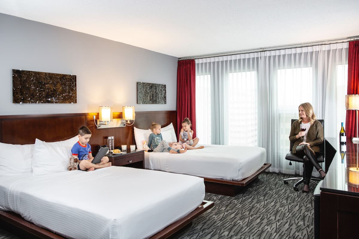 A hotel room with two beds, a desk, and a chair. There are two wall-mounted lamps, a window with red curtains, and artwork on the walls.
