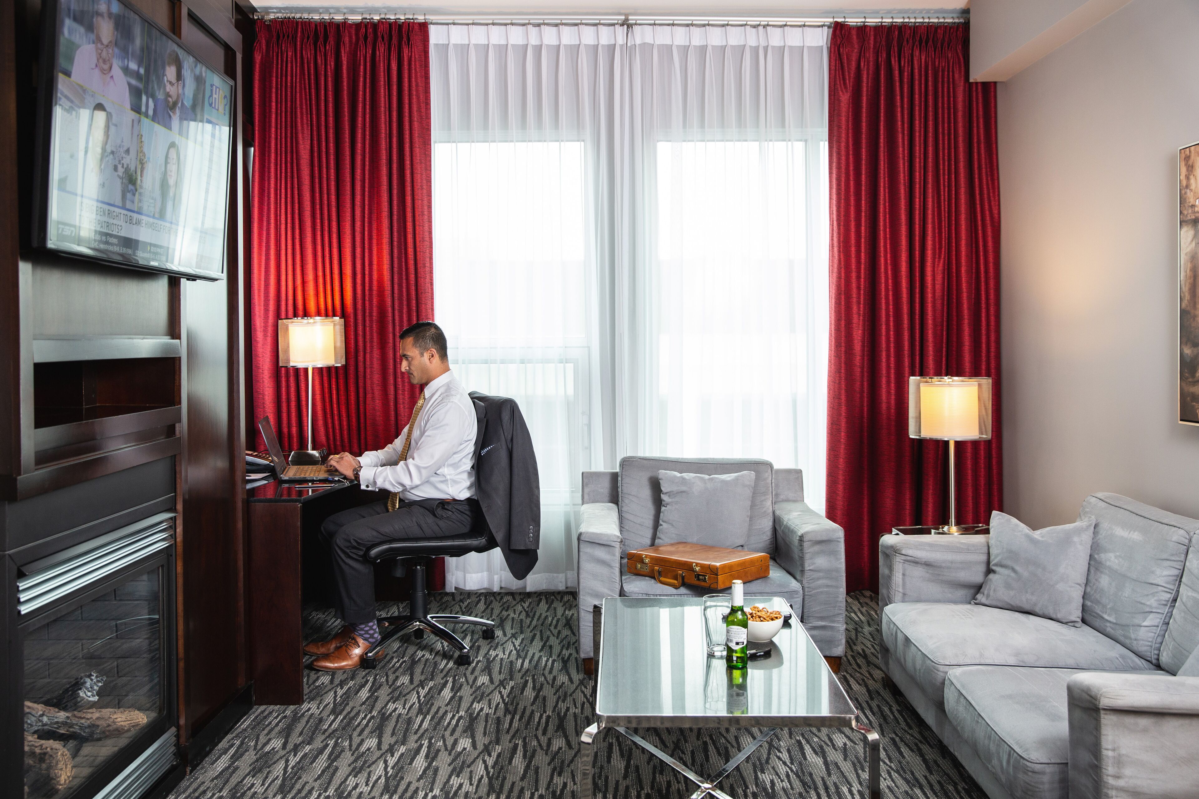 Une chambre d’hôtel avec un bureau, un ordinateur portable, une table basse en verre avec une bouteille et un bol, un canapé gris et des rideaux rouges.