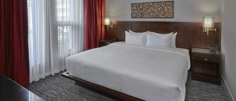 Habitación de hotel con una cama grande, una mesita de noche con lámpara, cabecero con panel decorativo y ventana con cortinas rojas.
