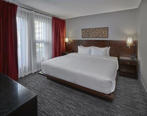 A hotel room with a large bed, a nightstand with a lamp, a headboard with a decorative panel, and a window with red curtains.