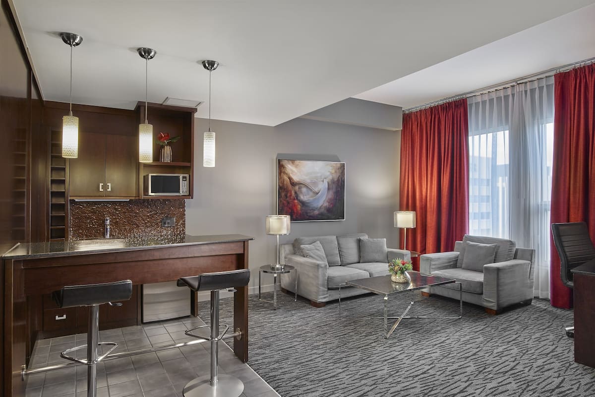 A modern hotel room with a kitchen area, two sofas, a coffee table, and a bar with stools.