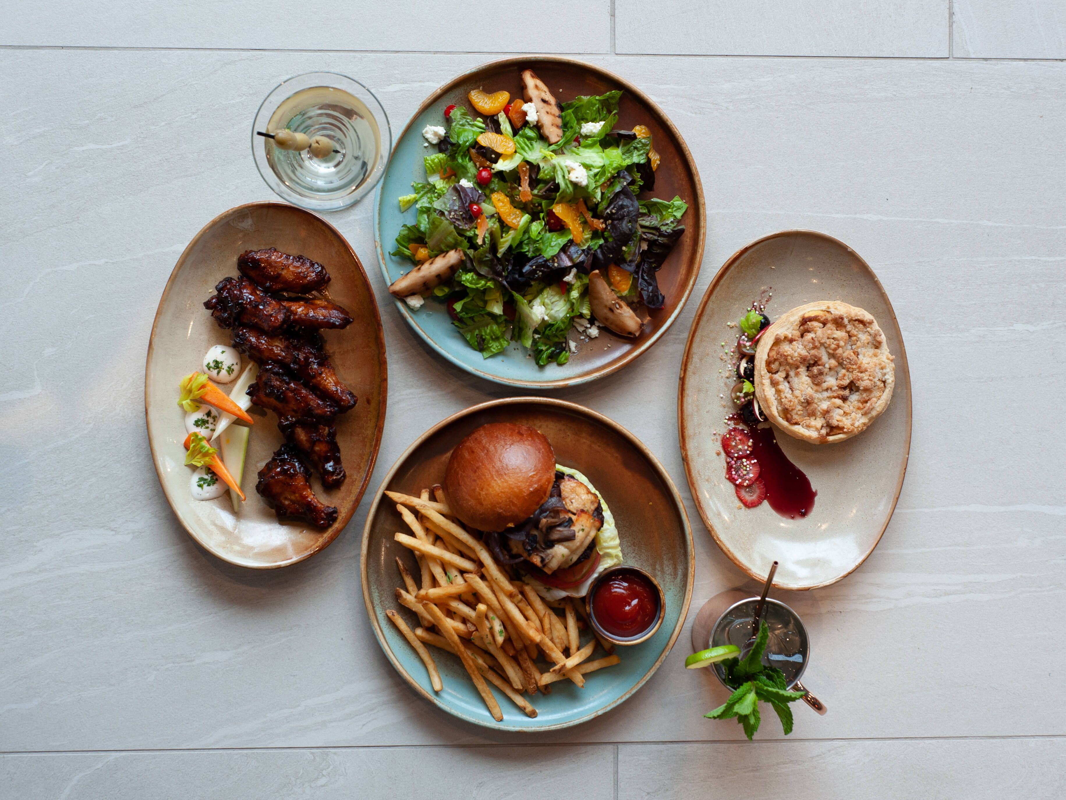 Uma mesa com diversos pratos, incluindo costelas, uma salada, um hambúrguer, batatas fritas e sobremesa.