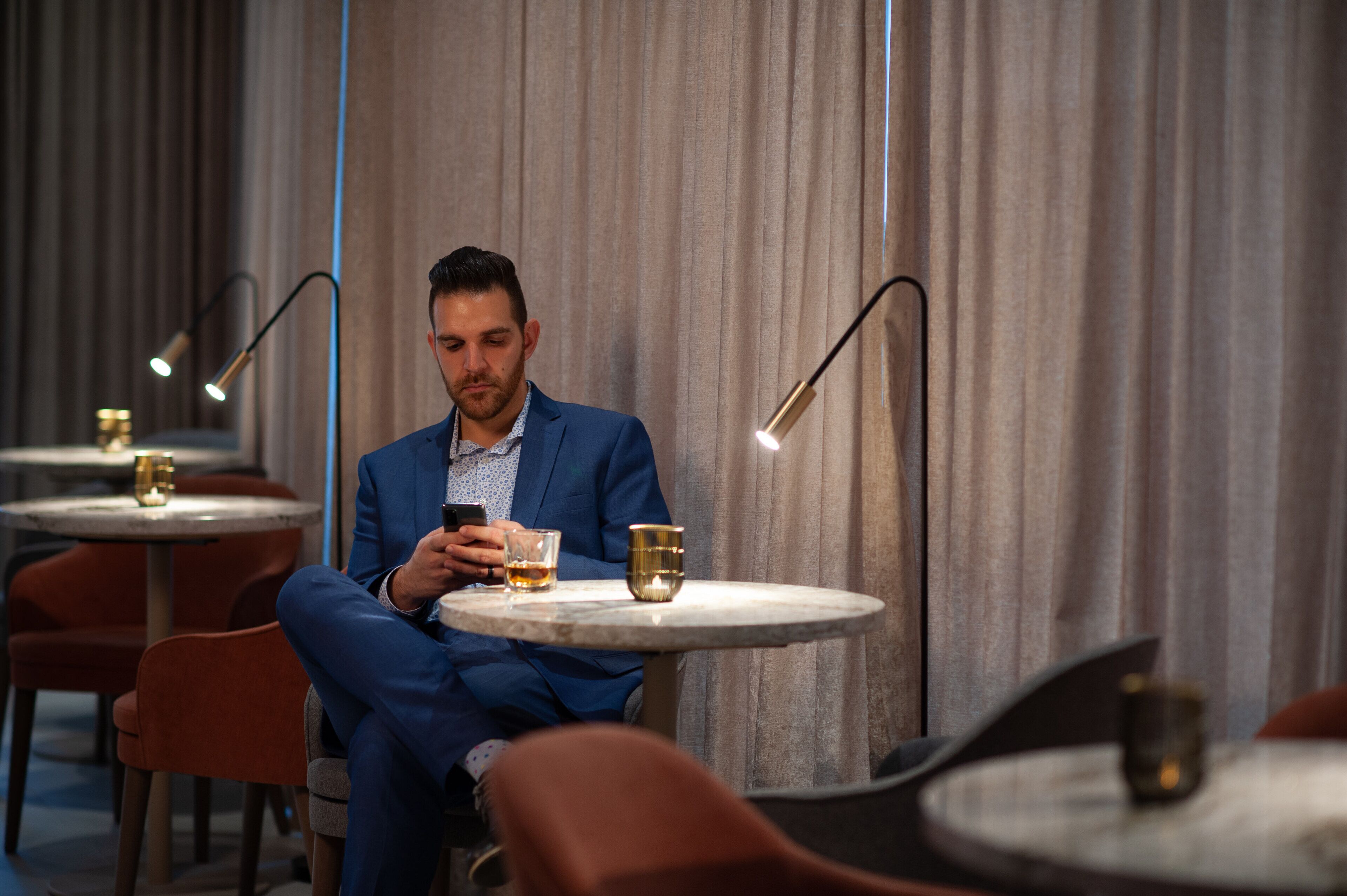 Un hombre con traje azul sentado a una mesa redonda con una bebida, en una habitación con cortinas e iluminación moderna.