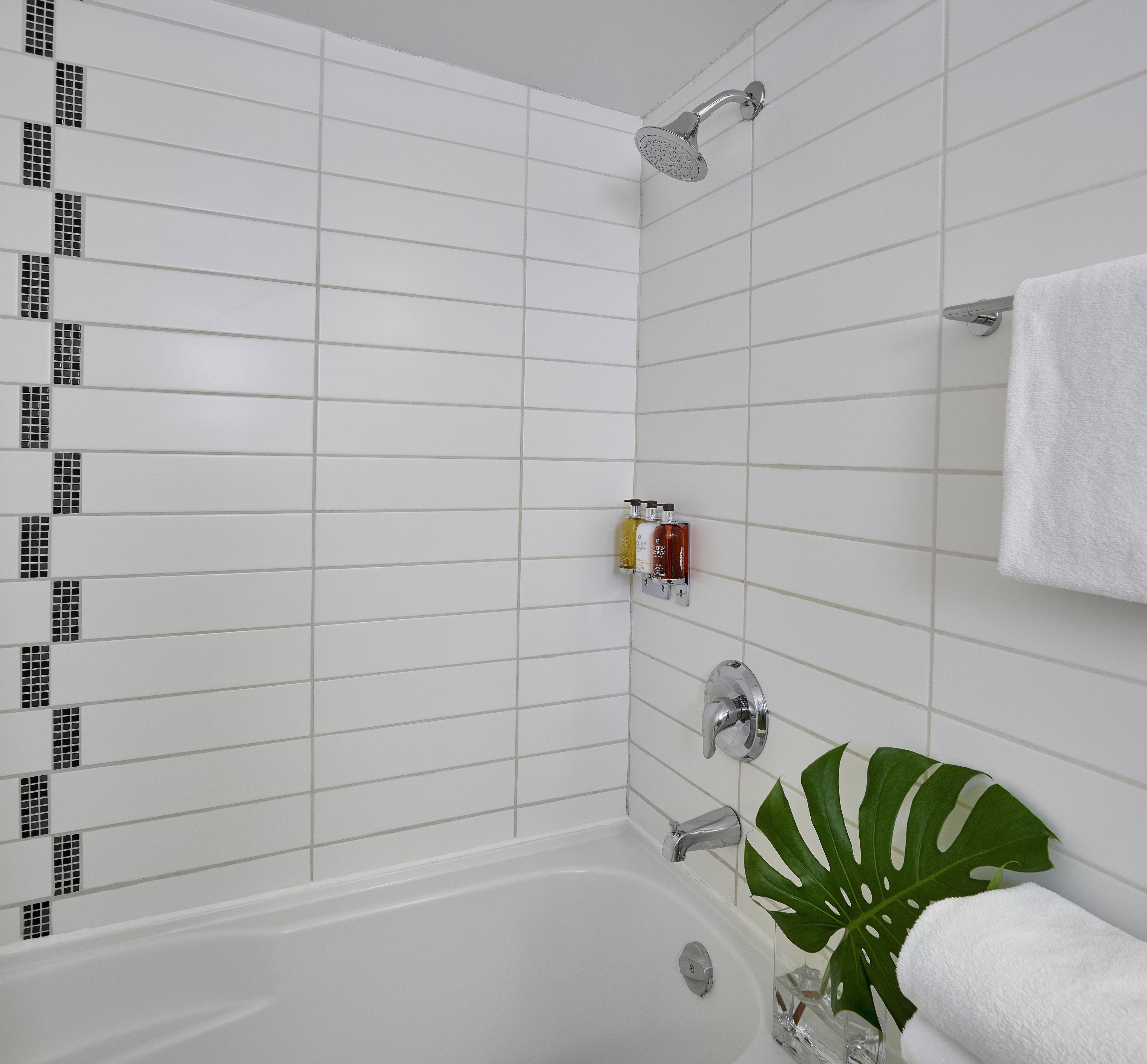 A modern bathroom with white tiled walls, a bathtub, a showerhead, and a potted plant.
