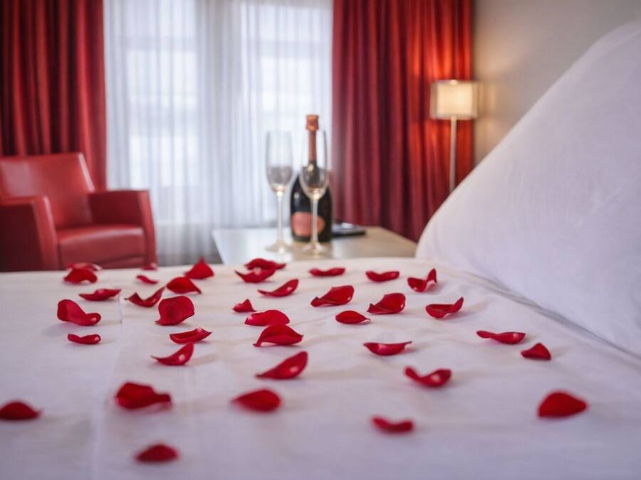 A hotel room with a bed covered in rose petals, a bottle of champagne, and a red chair.