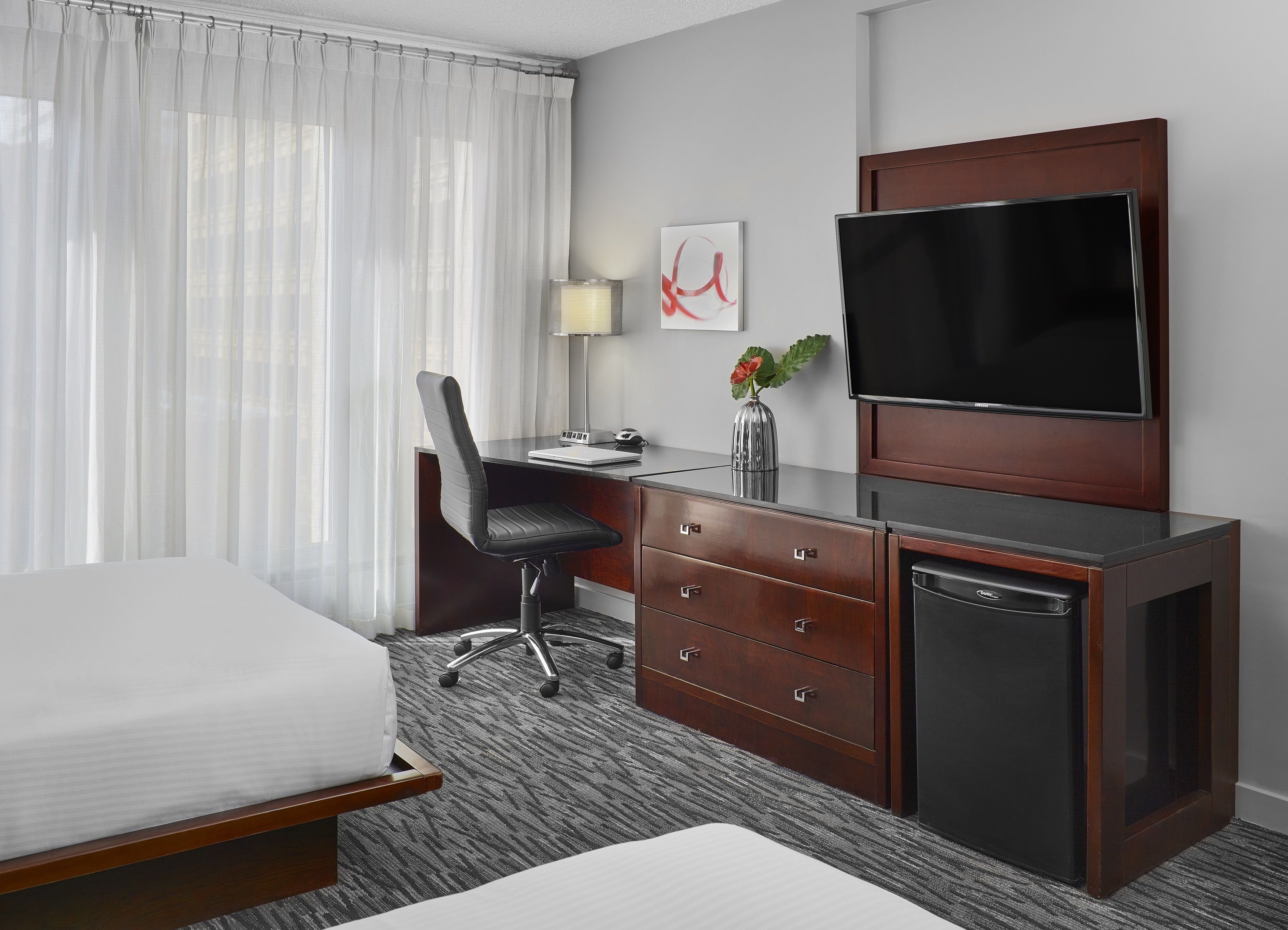 A hotel room with a bed, a desk with a chair, a television, a dresser, and a small refrigerator.