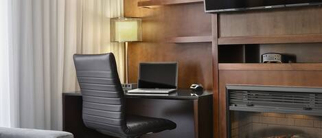 A modern hotel room with a black leather office chair, a laptop on a desk, a flat-screen TV, a fireplace with logs, and a vase with purple flowers on a shelf.