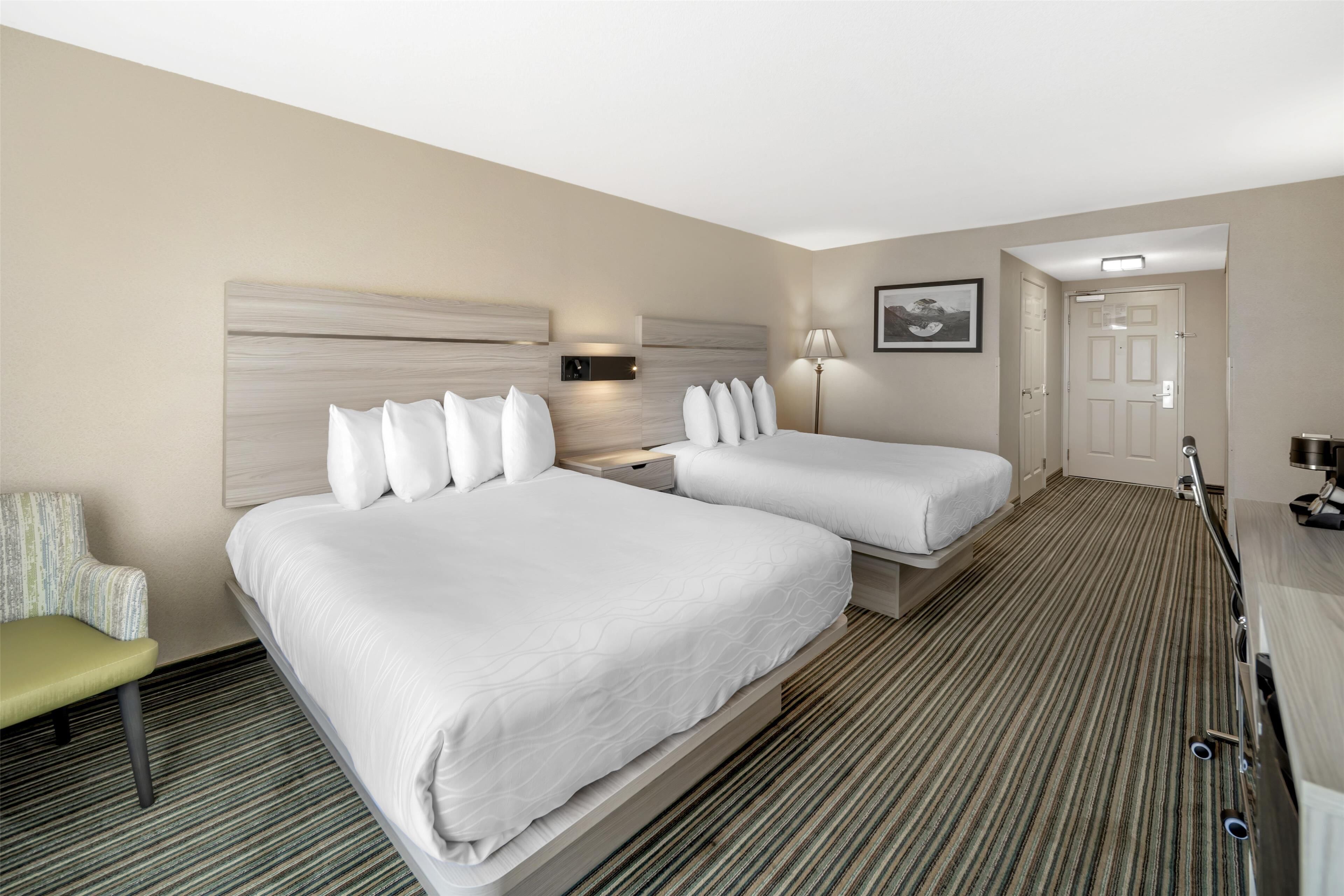A hotel room with two beds, a bedside table, a lamp, a chair, and a door.