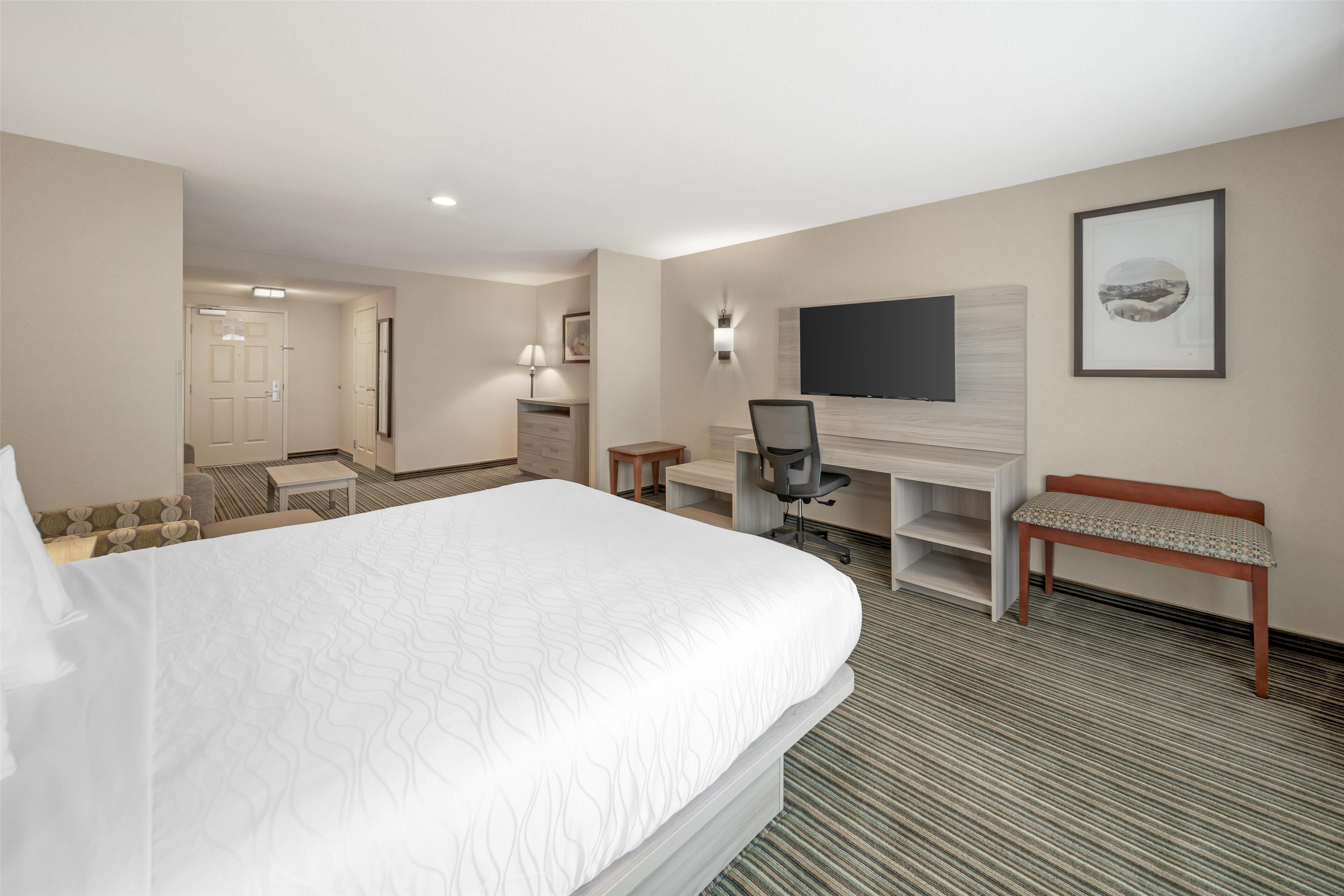 Ein Hotelzimmer mit einem großen Bett, einem Schreibtisch mit Stuhl, einem an der Wand befestigten Fernseher, einer Bank und einer Tür zu einem weiteren Raum.