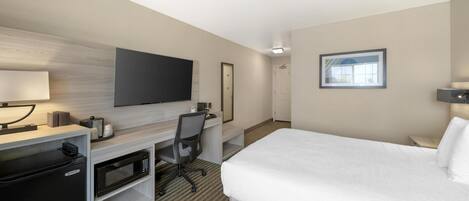 Habitación de hotel con cama, escritorio con computadora, microondas, refrigerador, televisor y un cuadro en la pared.
