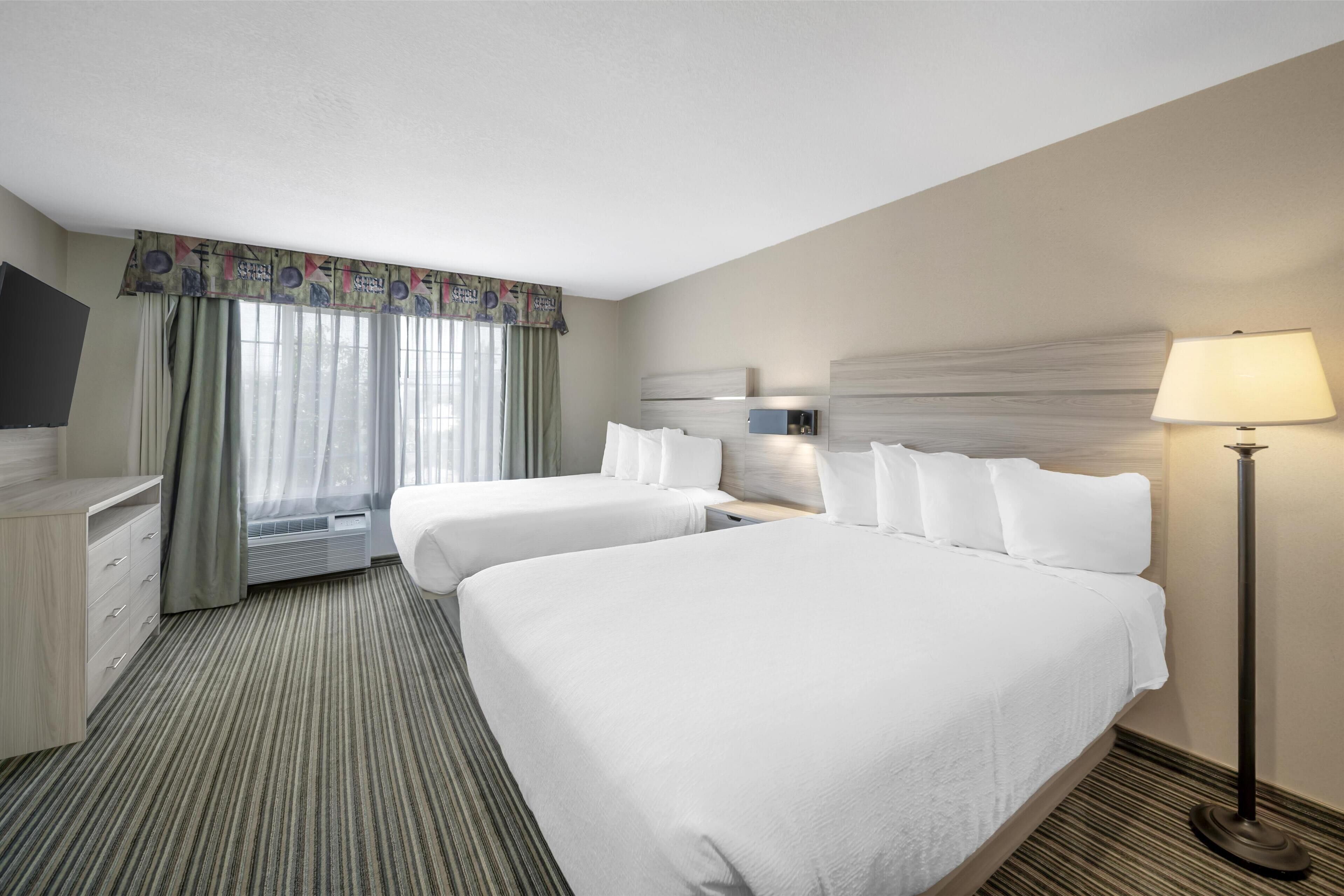 Ein Hotelzimmer mit zwei Betten, einem Schreibtisch, einem Flachbildfernseher, einem Fenster mit Vorhängen und einer Stehlampe.