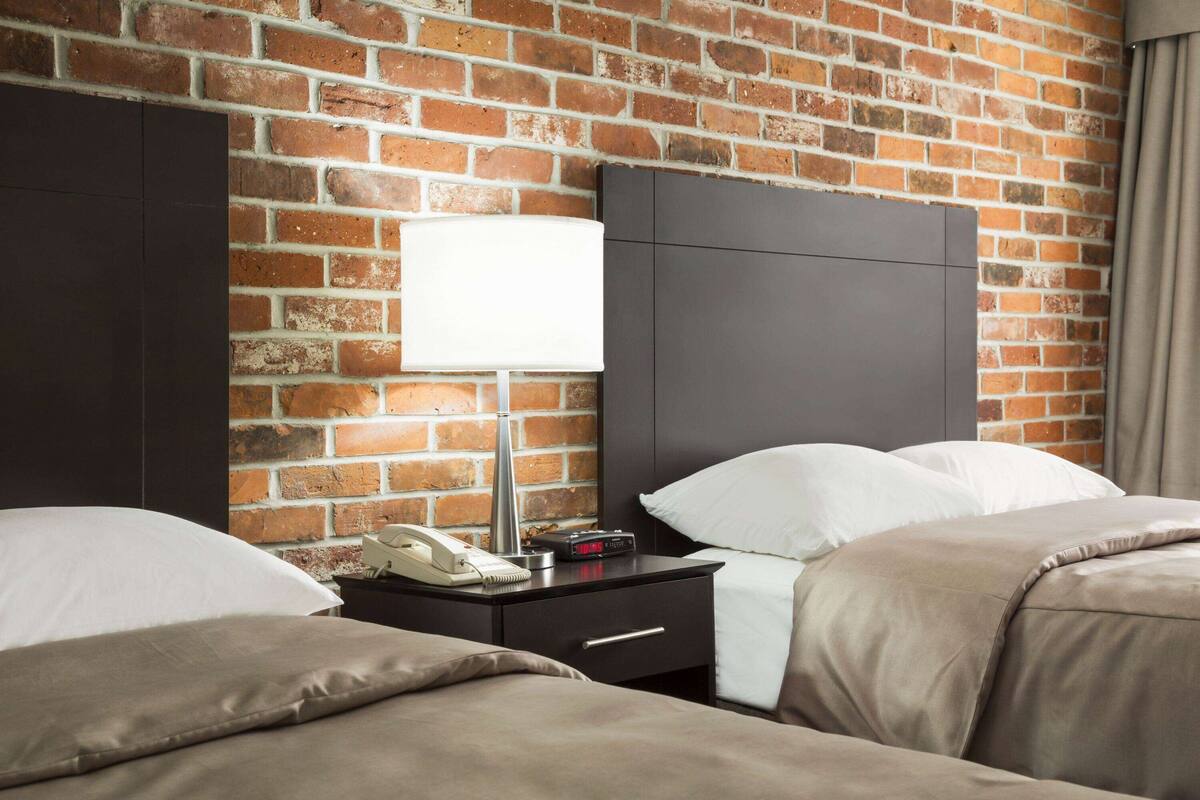 Hotellihuone, jossa on tiiliseinä, valkoisilla lakanoiden peitetty sänky, yöpöytä lampun kanssa ja puhelin.