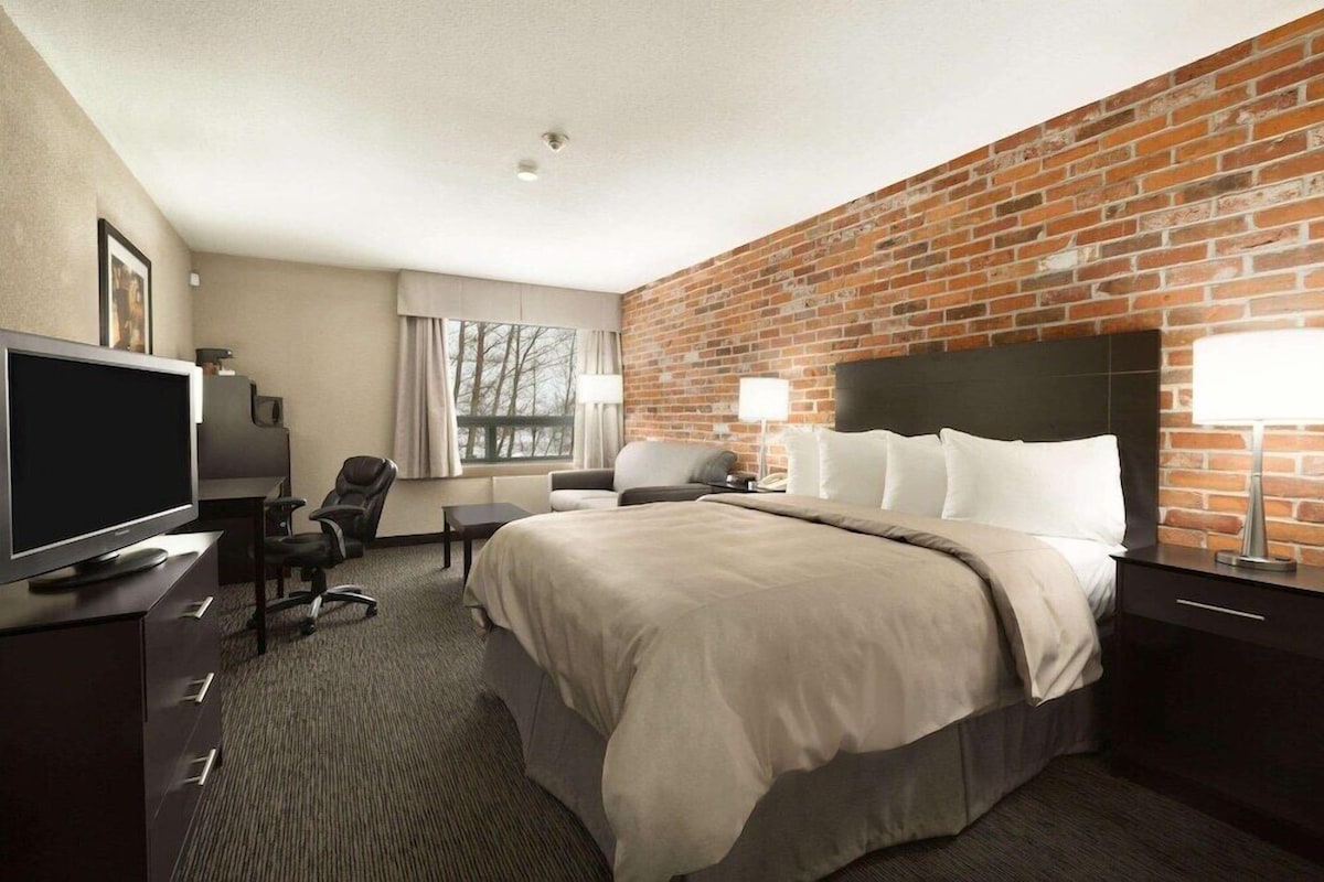 Ein Hotelzimmer mit Backsteinwand, einem Bett mit weißer Bettwäsche, einem Schreibtisch mit Fernseher und einem Sessel.