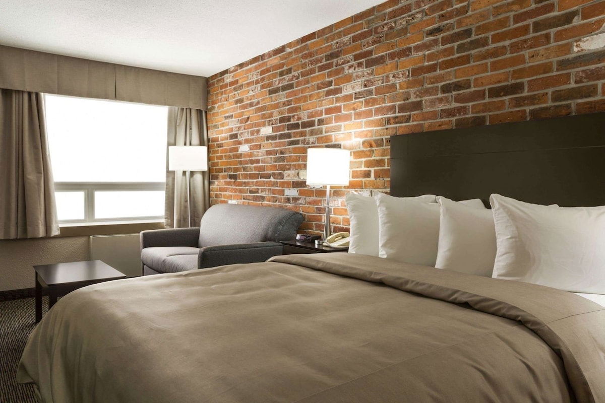 Ein Hotelzimmer mit Backsteinwand, einem Bett, einem Sessel, einem Nachttisch, einer Lampe und einem Fenster mit Vorhängen.
