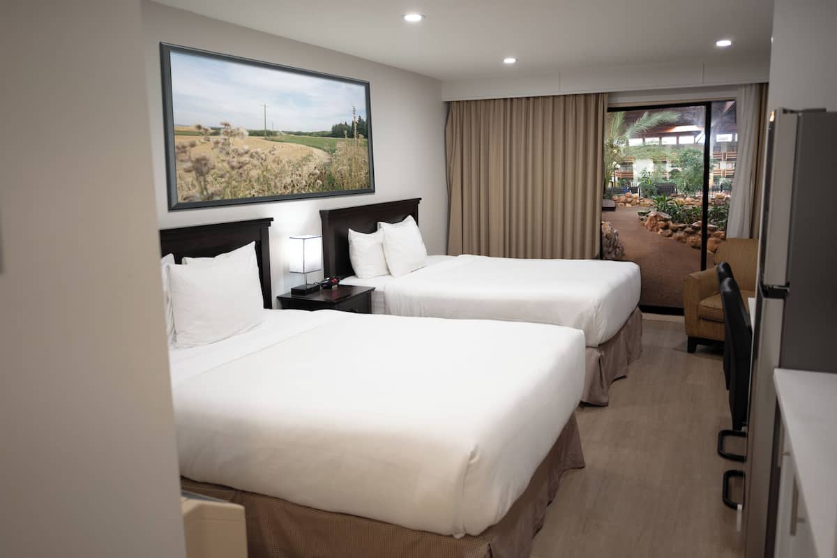 Ein Hotelzimmer mit zwei Betten, einem Schreibtisch, einem Sessel und einem großen Fenster mit Blick auf eine angelegte Grünfläche.