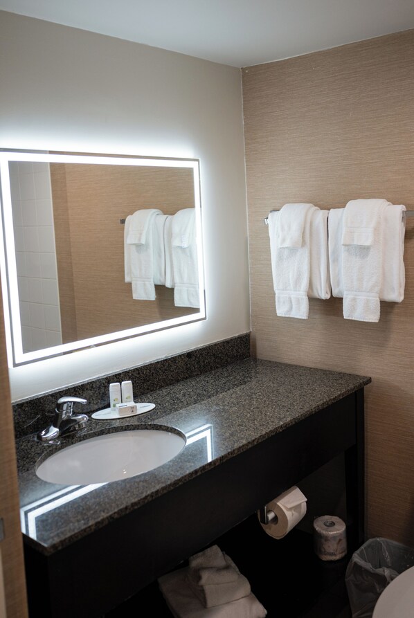 Un baño con lavamanos de encimera de granito, un espejo con iluminación integrada y un perchero con varias toallas blancas.