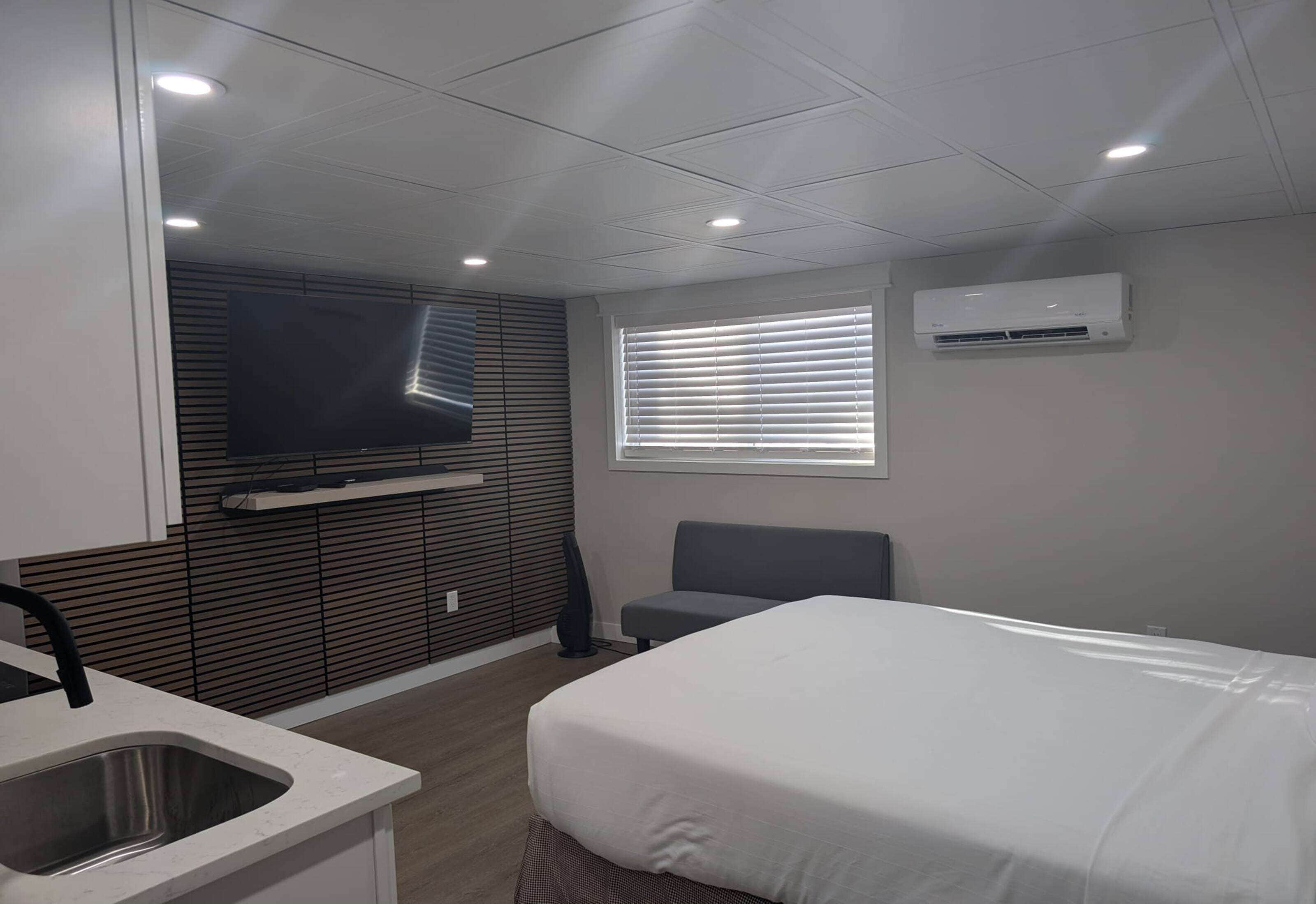 A modern hotel room with a bed, a TV mounted on the wall, a window with blinds, and an air conditioning unit.