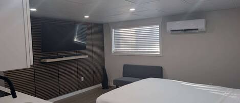 A modern hotel room with a bed, a TV mounted on the wall, a window with blinds, and an air conditioning unit.