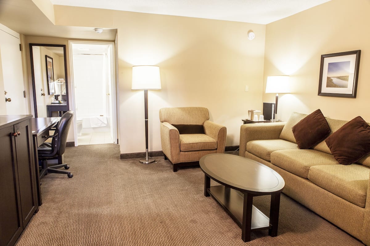 Ein Hotelzimmer mit Sofa, Ohrensessel und kleinem Tisch. Es gibt einen Schreibtisch mit Stuhl und Lampe. Der Boden ist mit Teppich ausgelegt und eine Tür führt in einen anderen Raum.