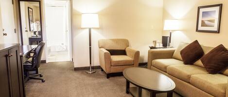 A hotel room with a sofa, armchair, and a small table. There is a desk with a chair and a lamp. The room has carpeted flooring and a door leading to another room.