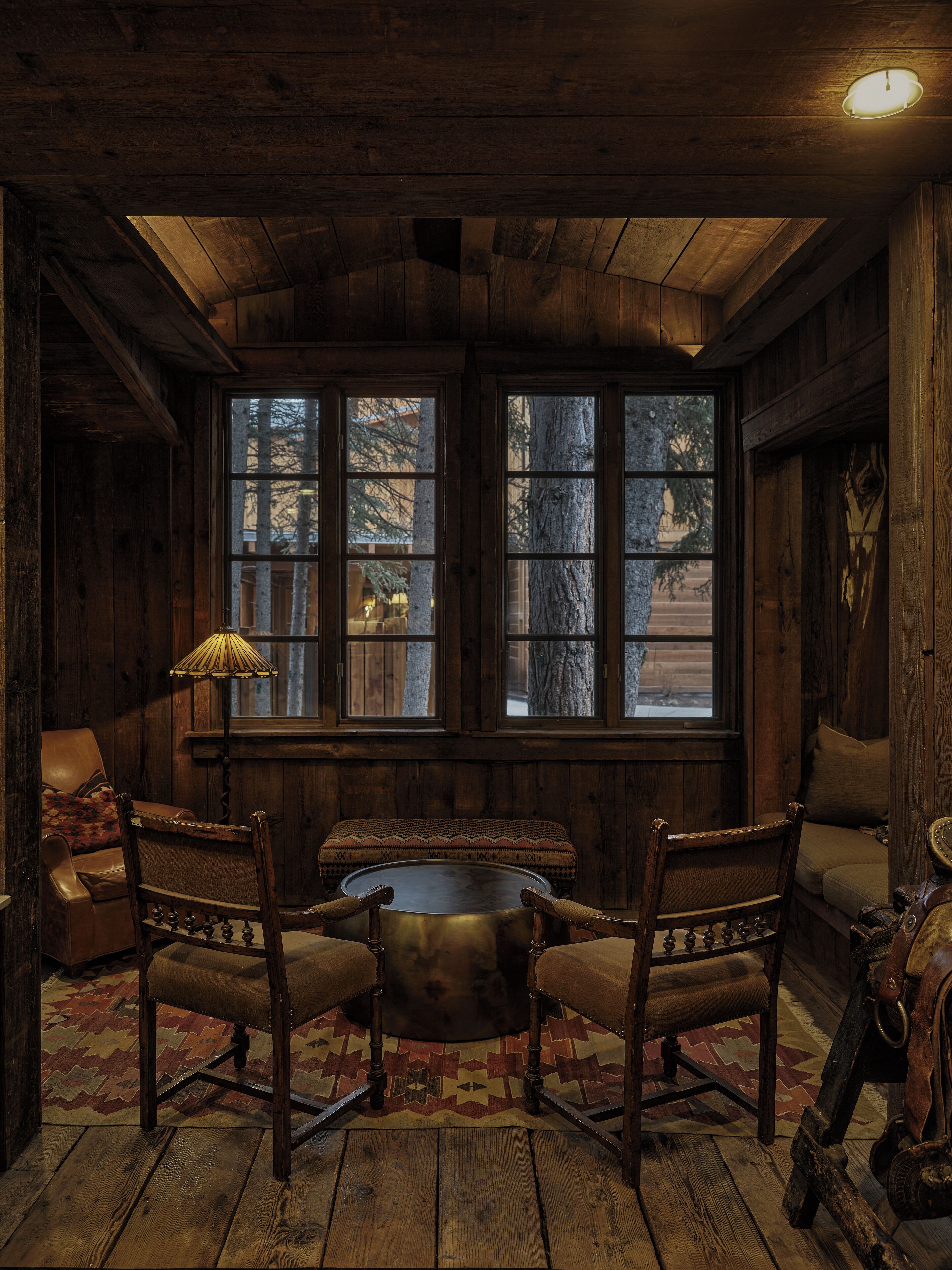 L’intérieur d’un chalet rustique, avec des murs en bois, une grande fenêtre donnant sur un paysage enneigé, une table ronde en bois et deux fauteuils en cuir.