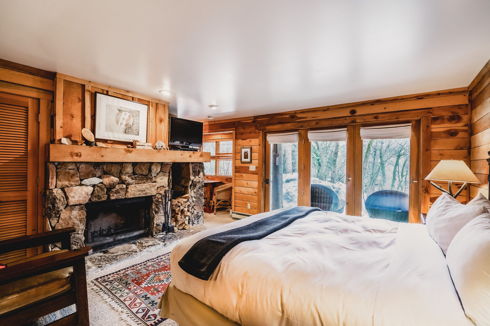 Un acogedor dormitorio de cabaña de madera con chimenea de piedra, una cama con manta y vistas al bosque a través de amplios ventanales.