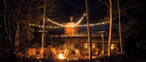 A rustic cabin with a fire, surrounded by trees and string lights.