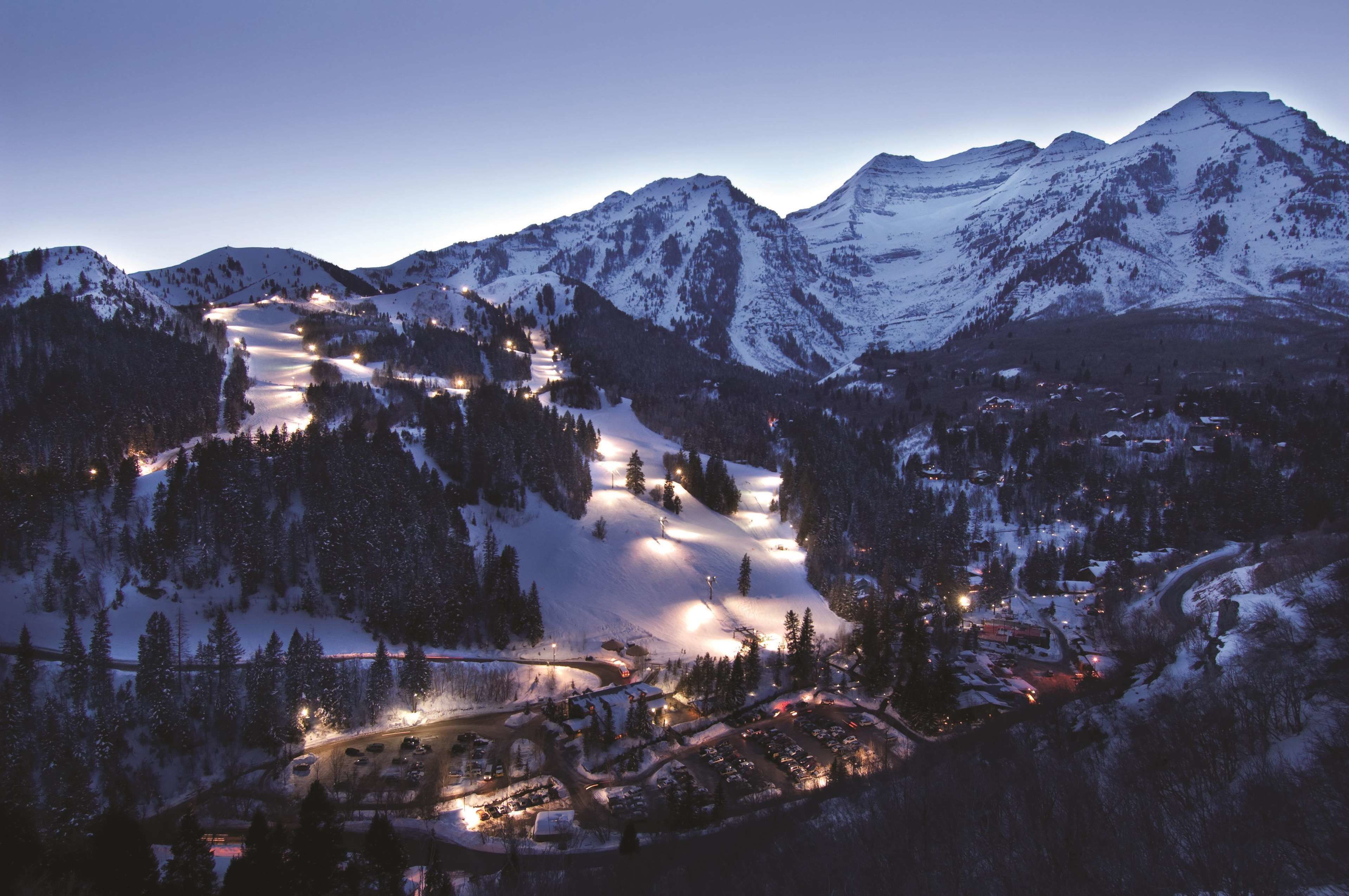 A snow-covered mountain resort at dusk with illuminated ski slopes and a small town below.