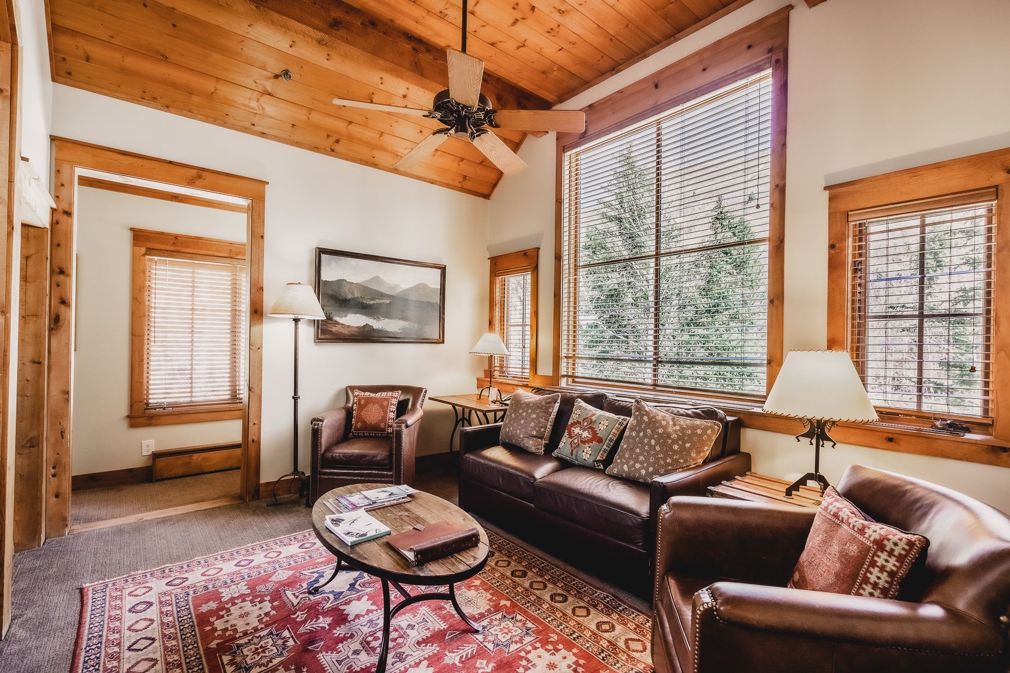 A cozy living room with a wooden ceiling, a brown leather sofa, a patterned rug, and large windows with blinds.