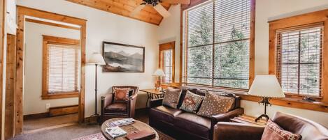 A cozy living room with a wooden ceiling, a brown leather sofa, a patterned rug, and large windows with blinds.