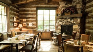 A rustic dining area with wooden tables and chairs, a stone fireplace, and framed pictures on the walls.