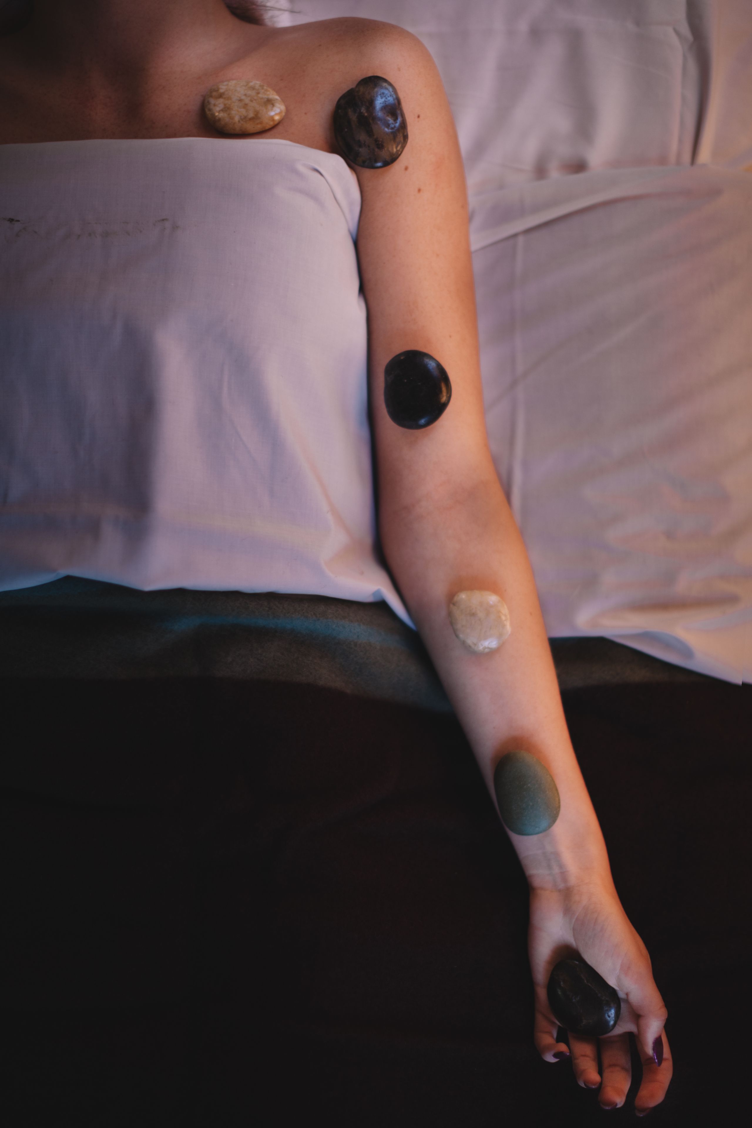A person lying on a bed with a white sheet, wearing a white top, and having three stones placed on their arm.
