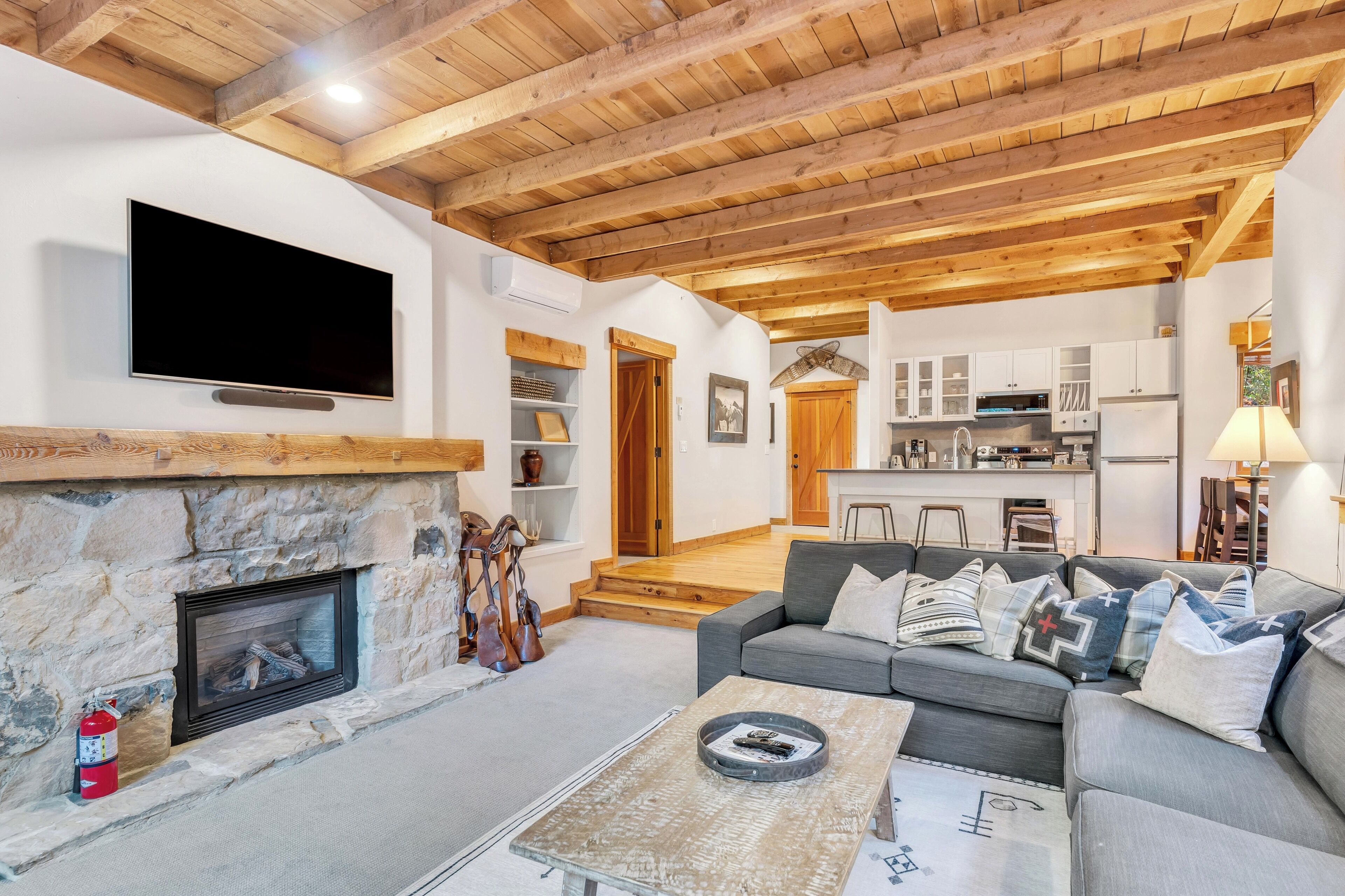 A spacious living area with a stone fireplace, a flat-screen TV mounted on the wall, and a modern kitchen in the background.