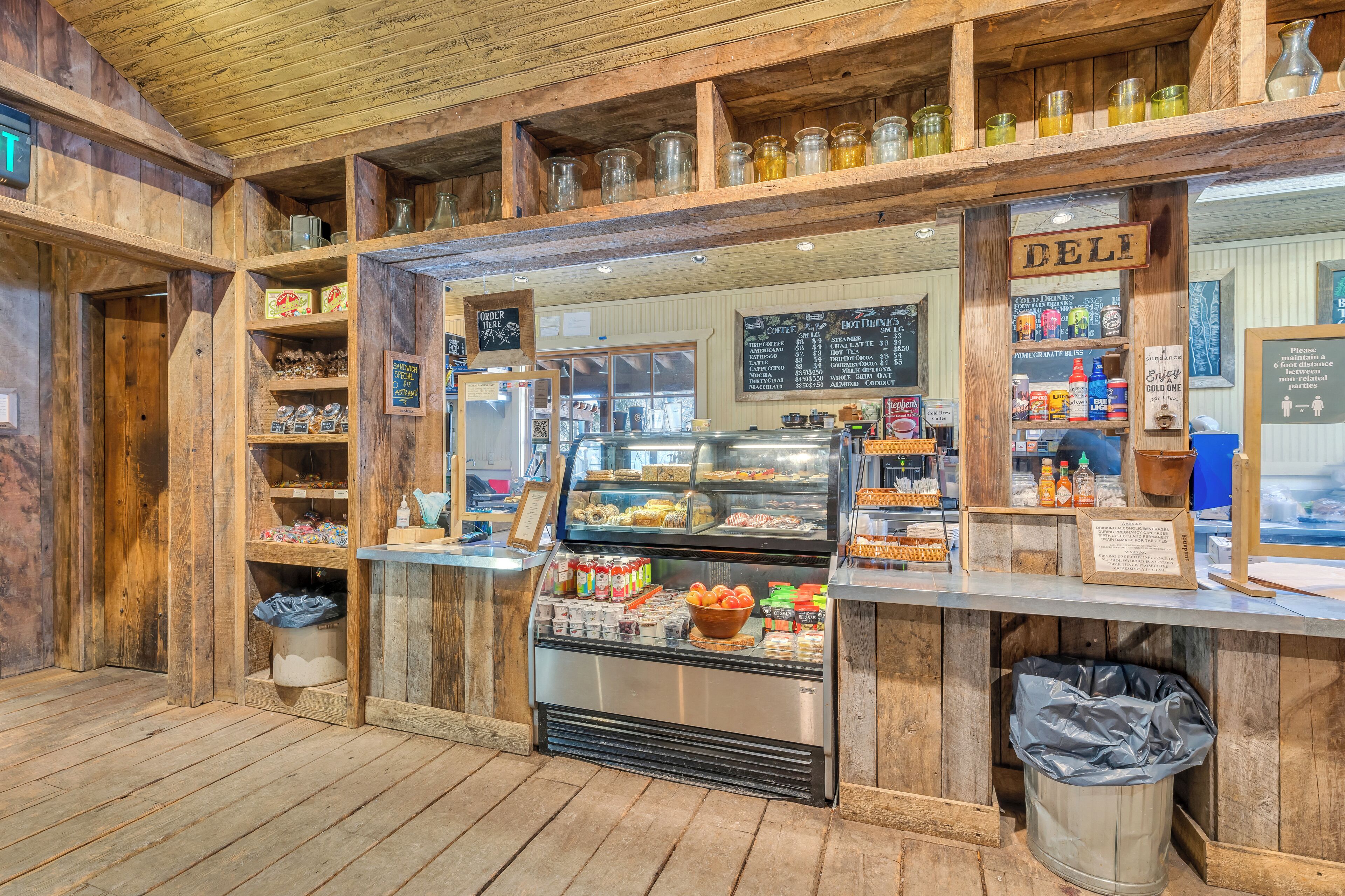 A deli counter with a glass display case, shelves with jars and bottles, and a menu board.