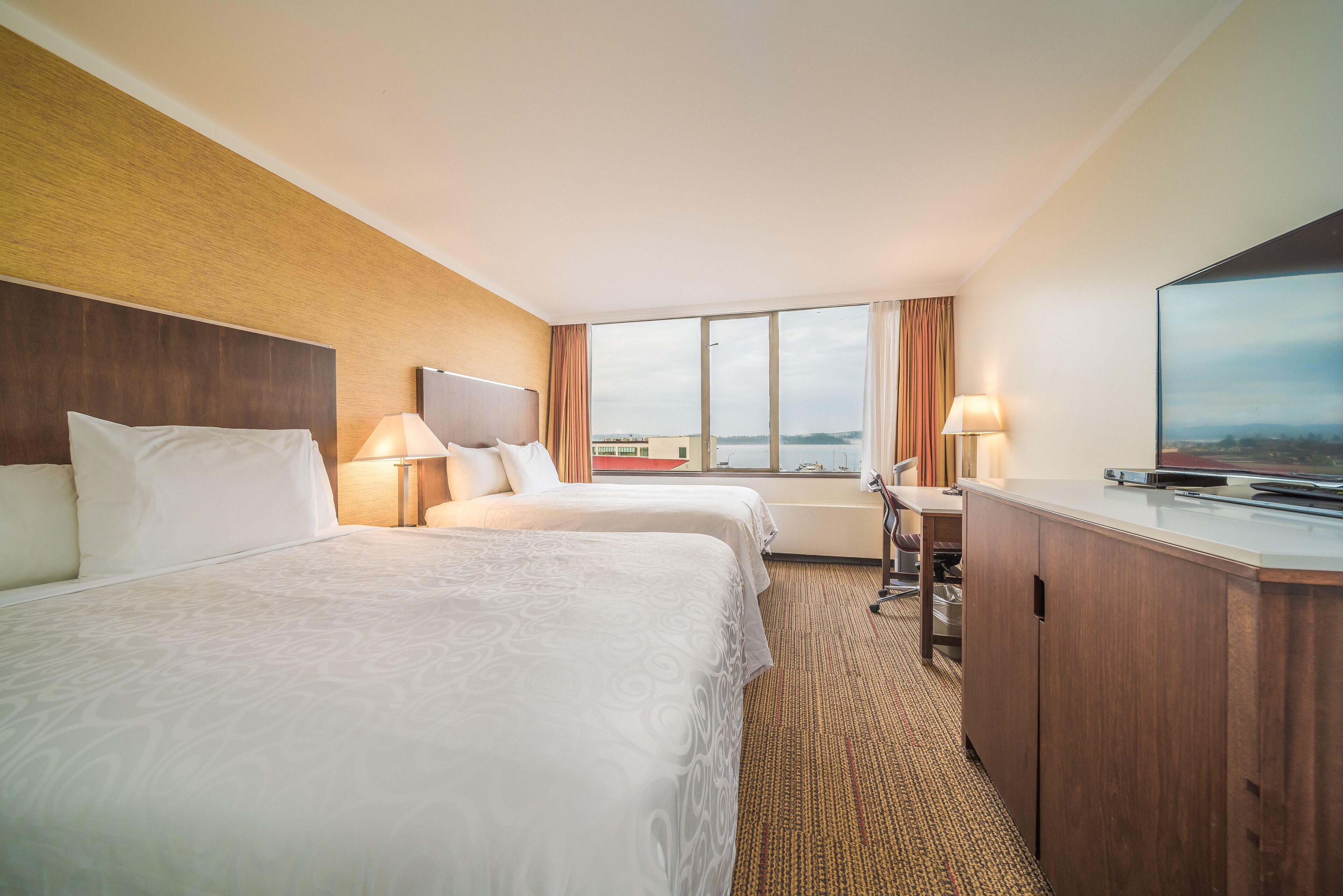 A hotel room with two beds, a desk, a television, and a view of the city.