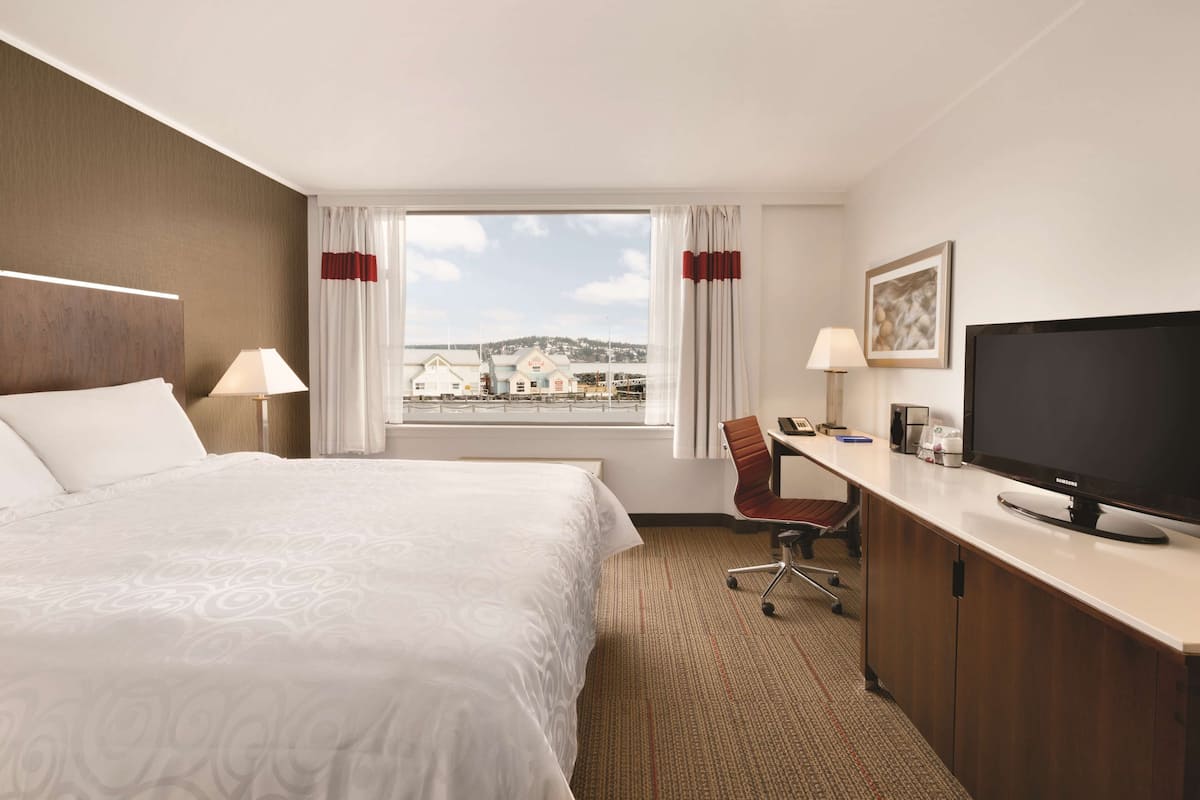 Habitación de hotel con una cama grande, un escritorio con una computadora, un televisor y vistas a edificios a través del ventanal.