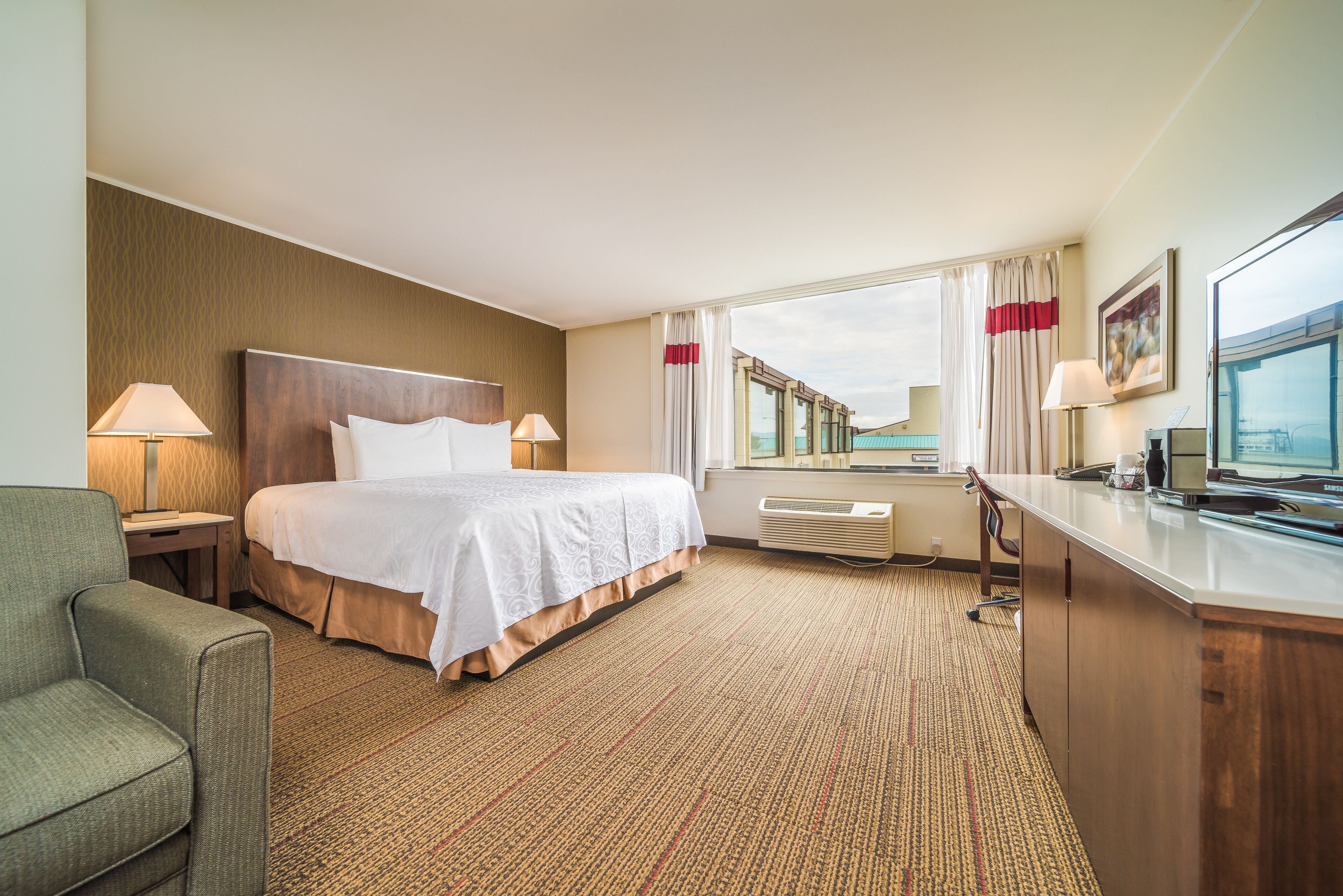 A hotel room with a large bed, a desk with a computer, a chair, and a window with curtains.