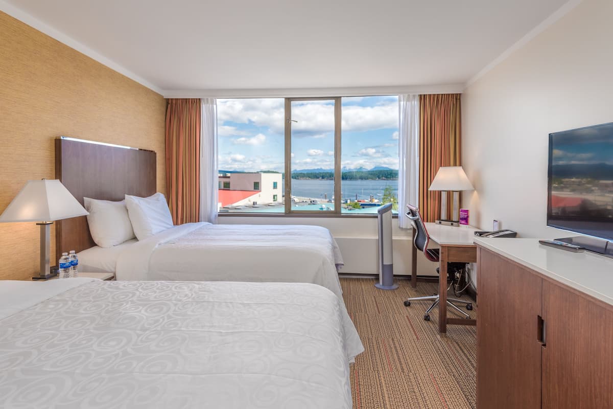 Habitación de hotel con dos camas, un escritorio, una silla, un televisor y vistas al agua y edificios.