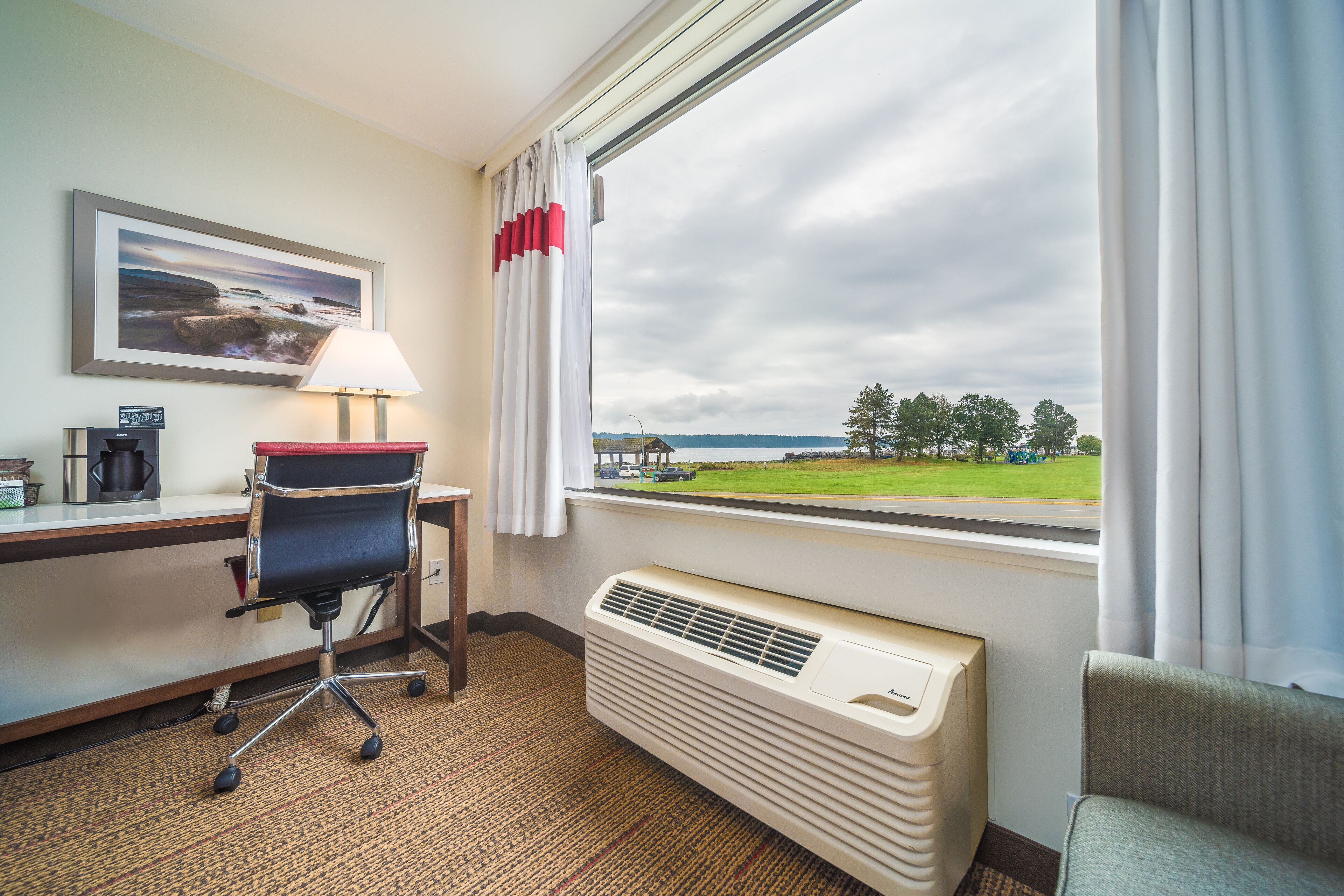A hotel room with a desk, chair, lamp, radiator, and a large window with a view of a green landscape and a building.