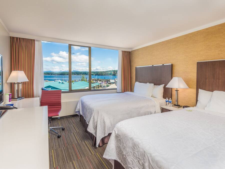 Habitación de hotel con dos camas, un escritorio y un amplio ventanal con vistas a un lago y montañas.