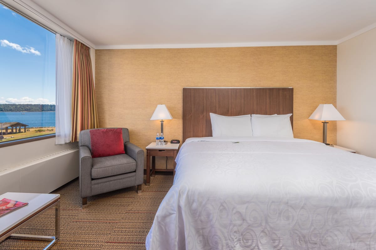 Habitación de hotel con una cama grande, dos lámparas de noche, una silla, una mesita con una revista y vistas al mar.