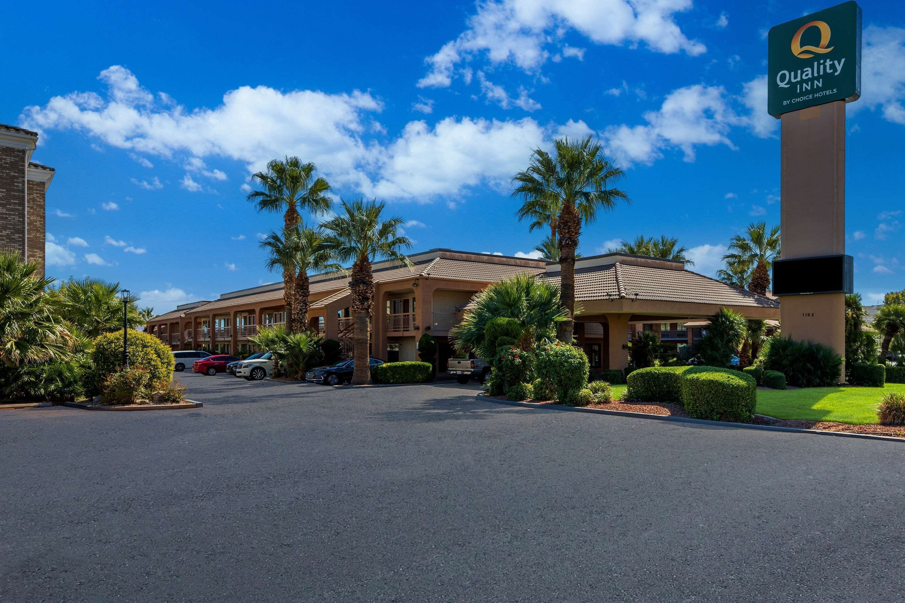 A hotel with a sign reading "Quality Inn by Choice Hotels," featuring a parking lot, palm trees, and a clear blue sky.