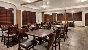A spacious dining area with multiple wooden tables and chairs, a stone accent wall, and a white door. - Quality Inn Saint George South Bluff (St. George)