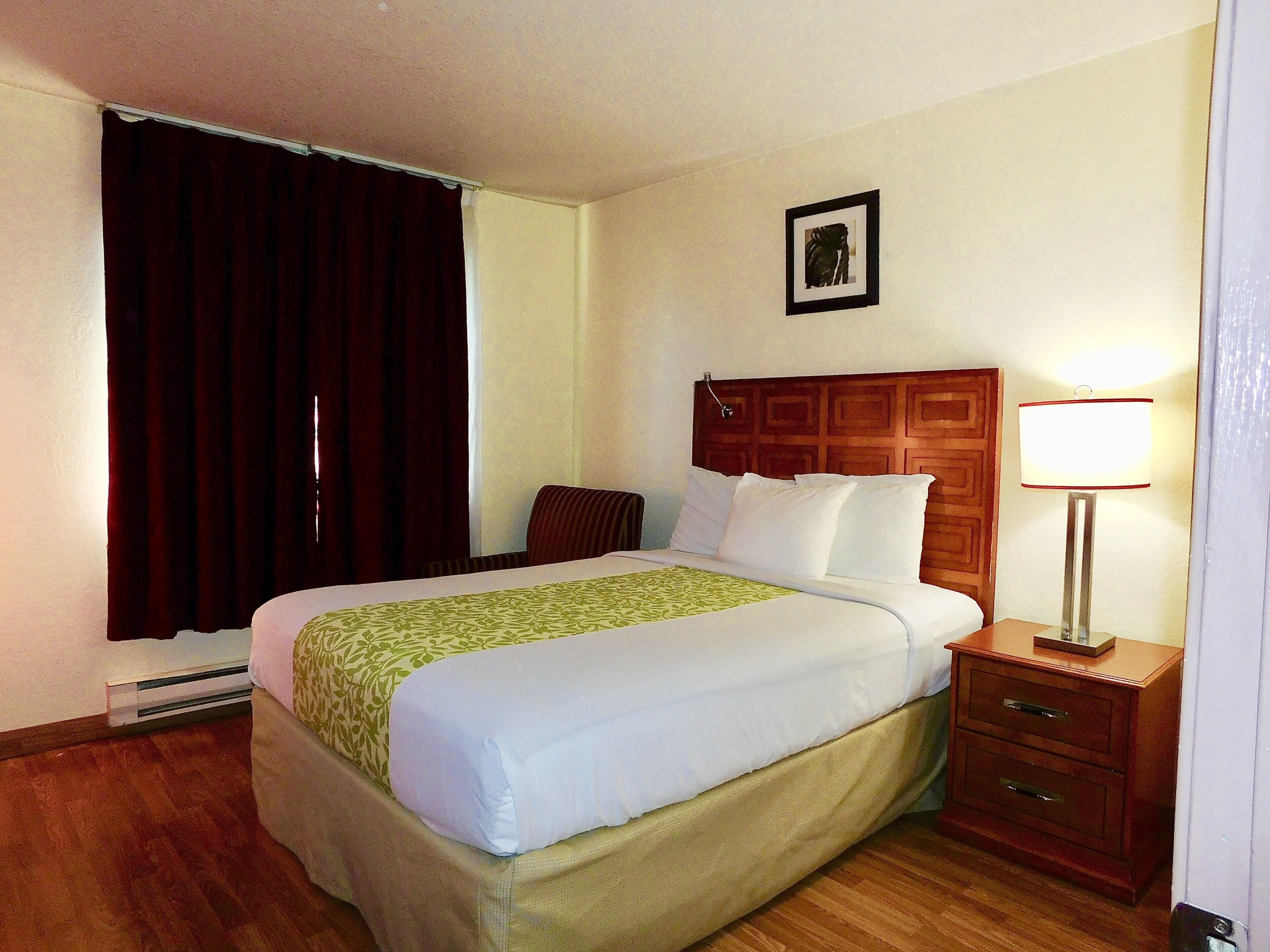 A hotel room with a bed, a nightstand, a lamp, a picture on the wall, and a window with curtains.