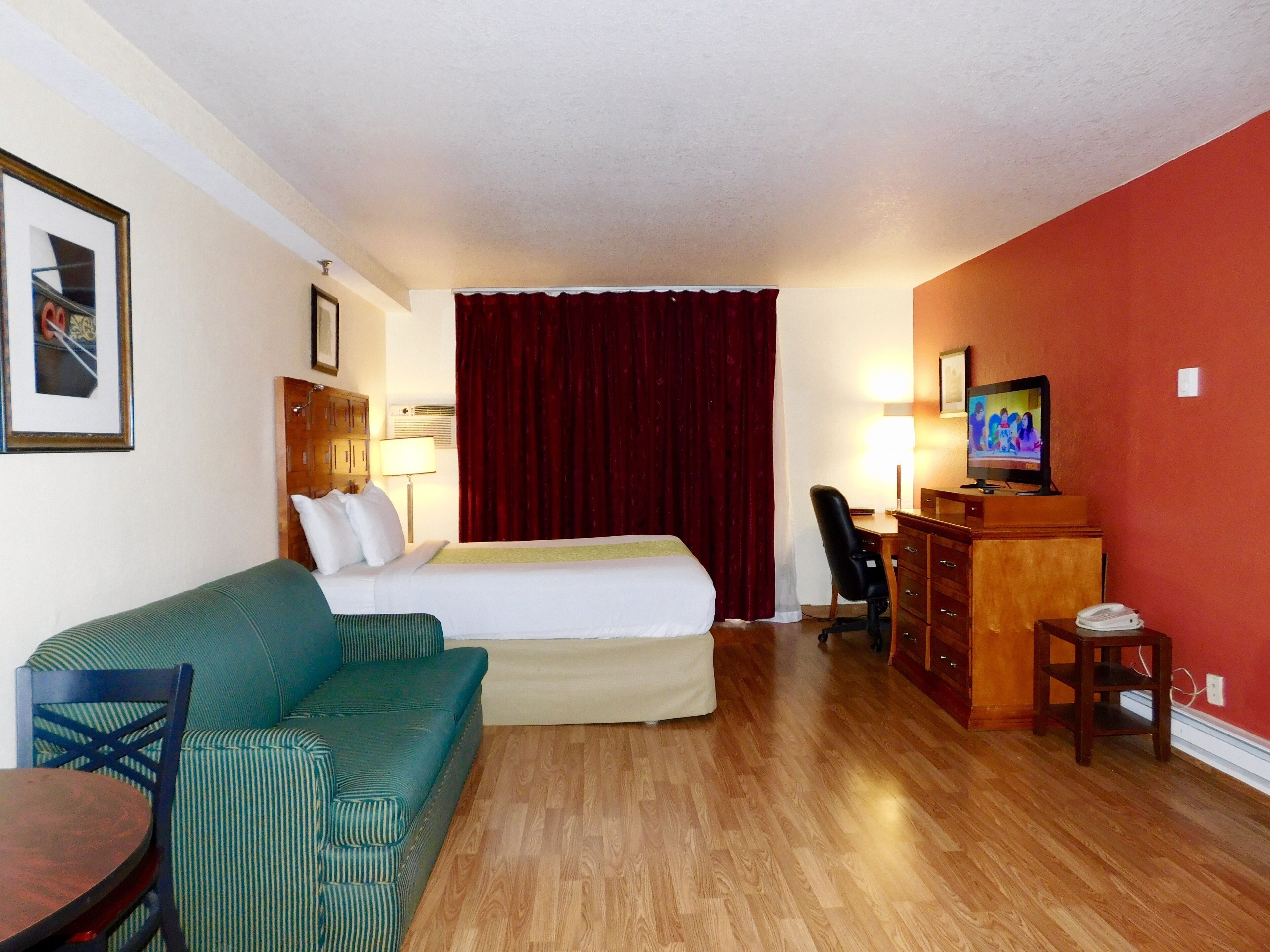 A hotel room with a bed, a green sofa, a wooden desk with a TV, and a small table.