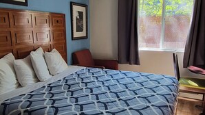 A bedroom with a bed, wooden headboard, two framed pictures, a window with curtains, and a chair.