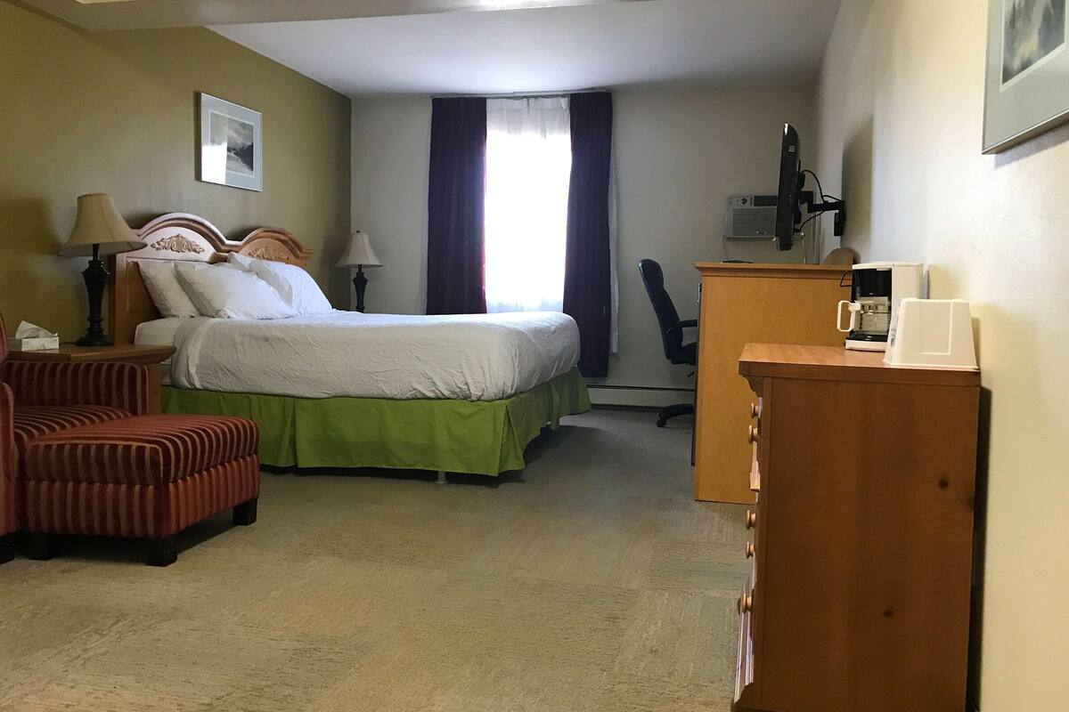 Une chambre d’hôtel avec un lit, un bureau, une chaise, une lampe, un tableau au mur et une fenêtre avec des rideaux.