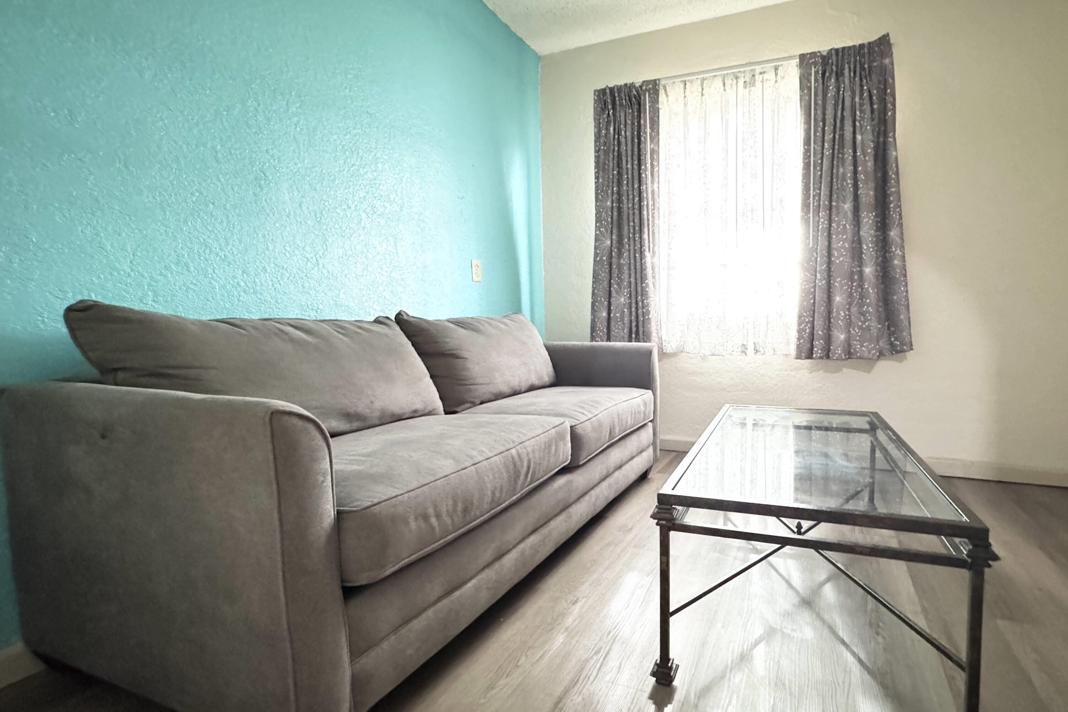 Un salon avec un canapé, une table basse en verre, une fenêtre avec des rideaux et un mur de couleur turquoise.