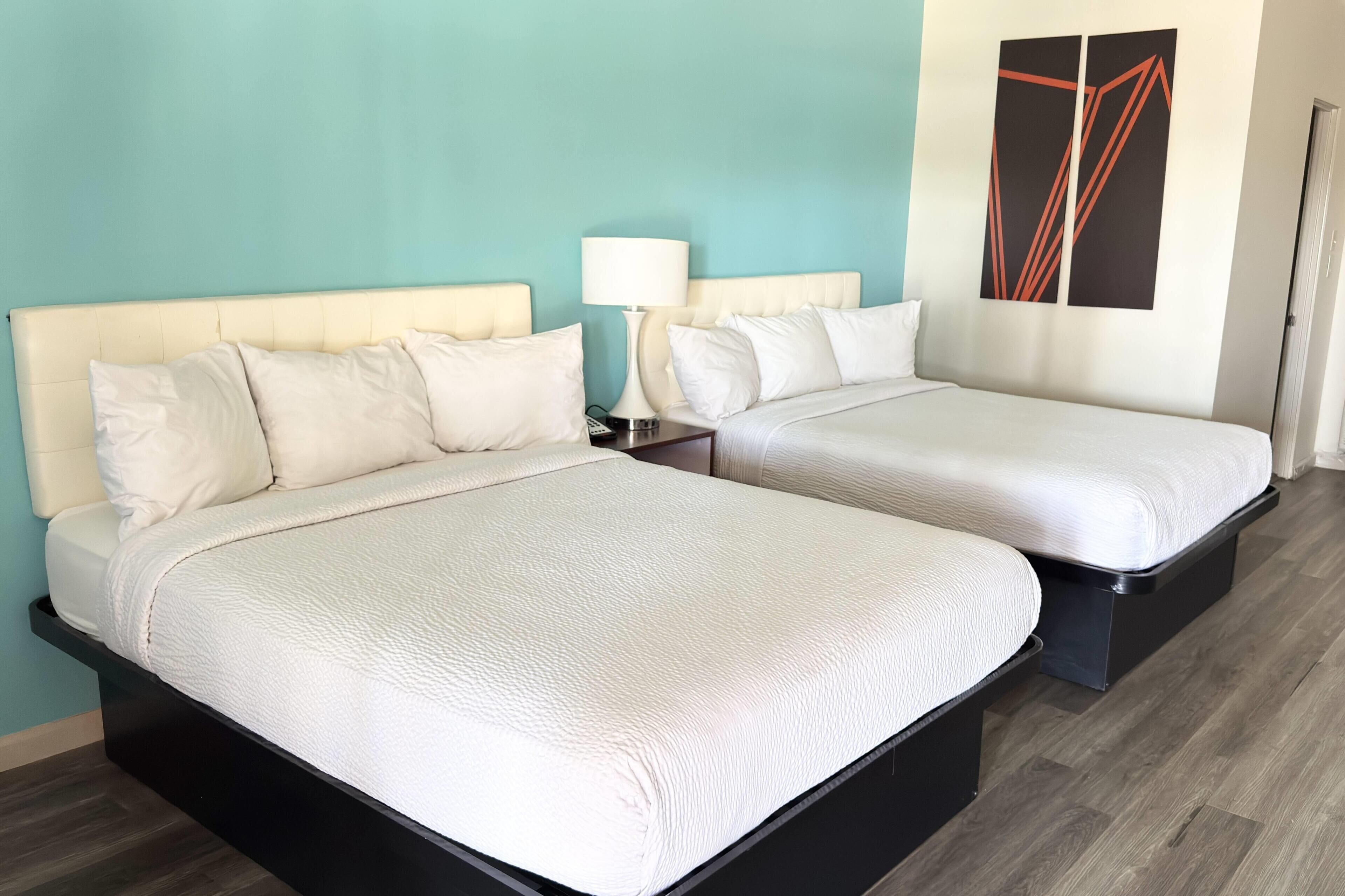 Quarto de hotel com duas camas, uma mesa de cabeceira com abajur e uma obra de arte na parede.